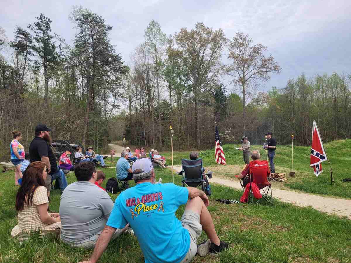 People sitting on grass, listening to two men standing with flags