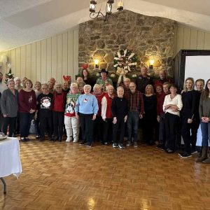 Several people standing in rows on side of room with stone fireplace and large screen