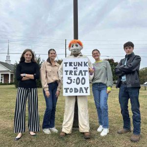Four teenagers standing next to masked figure with sign for trunk or treat.