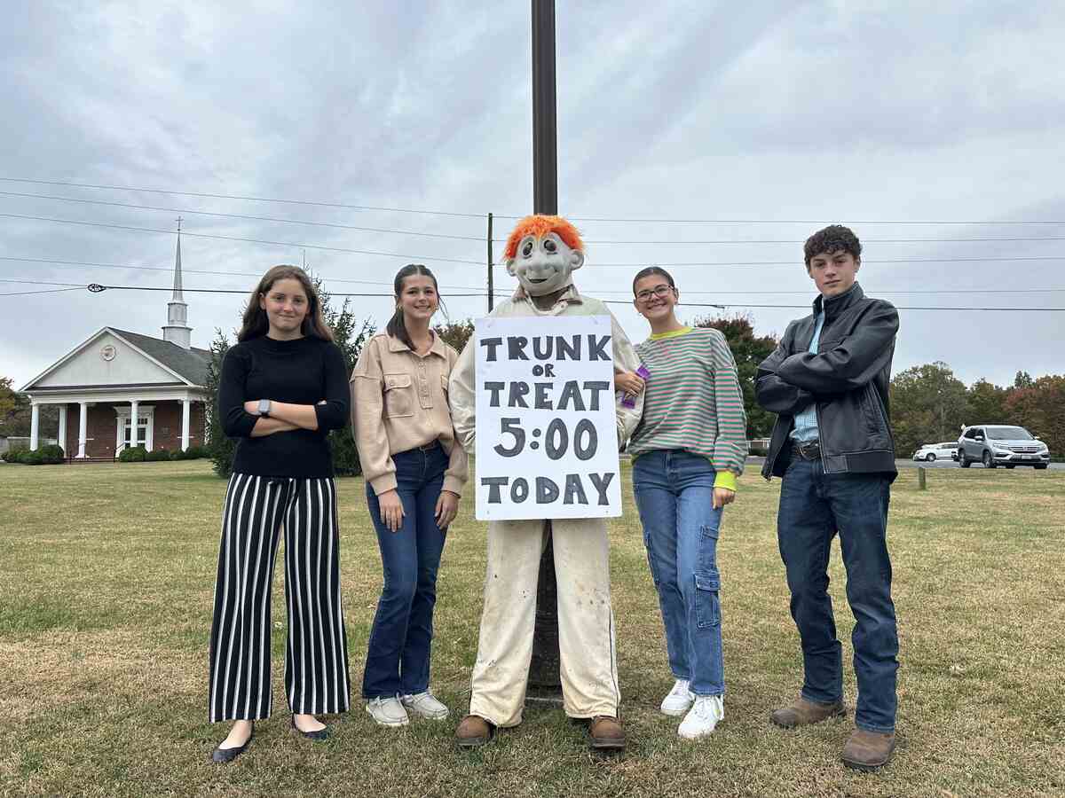 Four teenagers standing next to masked figure with sign for trunk or treat.